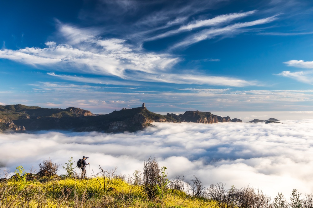 Wanderer fotografiert über einer Wolkendecke mit Blick auf Bergformationen und Roque Nublo, blauer Himmel mit Schleierwolken.